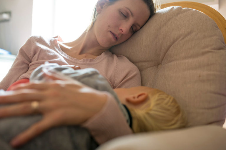 Tired young mother napping in a rocking chair with her baby sleeping in her lap.